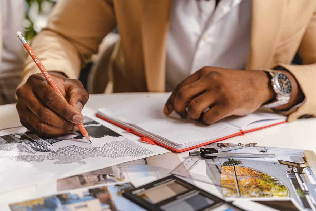 A person's hands sketch with a pencil over architectural drawings, building photographs and drafting tools on a desk.